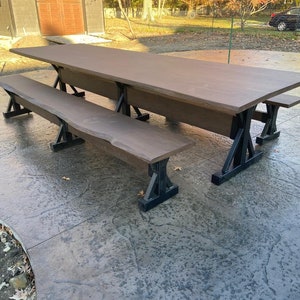 Pu&ograve; includere: Un grande tavolo da pranzo rettangolare con un piano in legno marrone liscio a bordo naturale, sostenuto da gambe in metallo nero a forma di X. Il set include due panche abbinate con gli stessi piani in legno marrone e gambe in metallo.