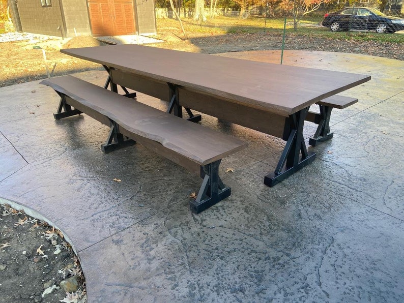 Pu&ograve; includere: Un grande tavolo da pranzo rettangolare con un piano in legno marrone liscio a bordo naturale, sostenuto da gambe in metallo nero a forma di X. Il set include due panche abbinate con gli stessi piani in legno marrone e gambe in metallo.