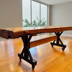 May include: A rectangular table featuring a thick, natural-edge wooden slab top with a dark brown finish. The base consists of black metal X-shaped supports connected by a horizontal wooden beam, all mounted on small black caster wheels for mobility.