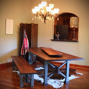 May include: A rustic wooden dining table with a live edge top and metal legs. The table is set with two benches and a cowhide rug. A chandelier hangs above the table.