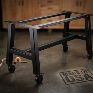 May include: A black metal table base with a rectangular frame and four caster wheels. The base features flat mounting plates at the top for a tabletop. A measuring tape is visible on the floor near the base, and the text MADE IN USA is present.