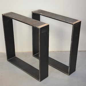 May include: Two black metal table legs with a square design. The legs are made of thick metal and have a rustic finish.