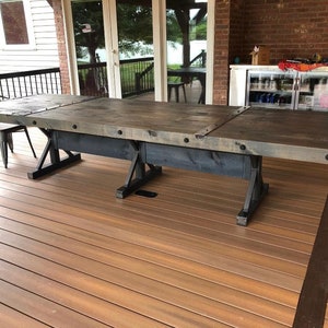 Pu&ograve; includere: Un lungo tavolo da pranzo rettangolare con un piano in legno tinto grigio scuro con venature visibili, accentuato da staffe angolari e bulloni in metallo nero. Il tavolo &egrave; sostenuto da una robusta base in metallo nero con design a X.