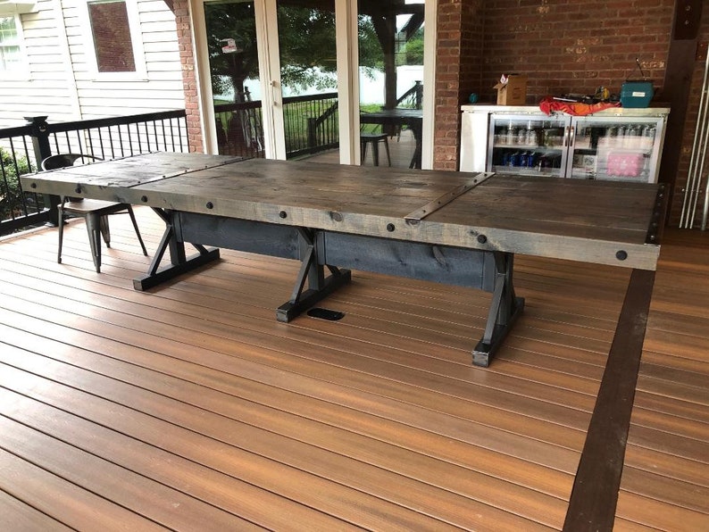 Pu&ograve; includere: Un lungo tavolo da pranzo rettangolare con un piano in legno tinto grigio scuro con venature visibili, accentuato da staffe angolari e bulloni in metallo nero. Il tavolo &egrave; sostenuto da una robusta base in metallo nero con design a X.