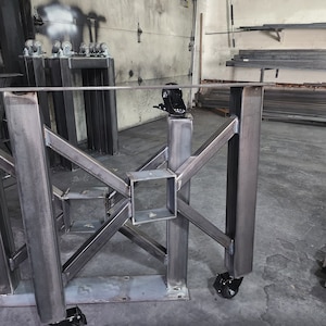 May include: A metal table frame with a black powder coat finish. The frame has a square design with multiple cross beams and is supported by four wheels. The table frame is in a garage setting.