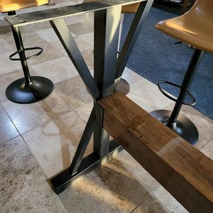 May include: A black metal table base with a wooden top. The base has a V-shaped design with four legs. The table is in a room with tile flooring.