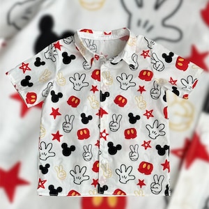 May include: White button-down shirt with a short sleeve featuring a repeating pattern of red, black, and yellow Mickey Mouse ears, hands, and stars.