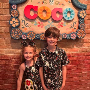 May include: Two children, a boy and a girl, stand in front of a wooden sign that reads "Plaza de Coco". The girl is wearing a black dress with a white skull and floral design. The boy is wearing a black short-sleeved shirt with a colorful print of characters from the movie Coco.