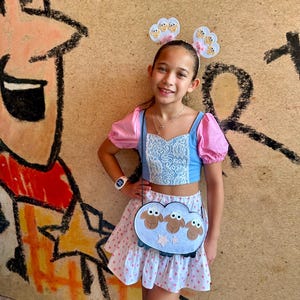 May include: A young person wearing a blue and pink crop top with a white lace bodice, a white and pink polka dot skirt, and a cloud-shaped purse with sheep faces. The outfit is accessorized with a headband with cloud and sheep details.