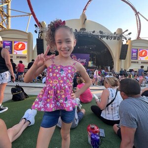 May include: A young girl with curly brown hair wearing a pink and purple sleeveless top with a cartoon print and blue denim shorts. She is smiling and making a peace sign with her hands. The girl is standing in front of a stage with a large screen behind her. The screen displays the words "Gumbo Gras".