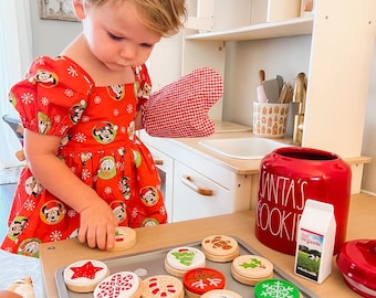 May include: A child in a red and white dress with a Mickey Mouse pattern is playing with a set of wooden Christmas cookie play food. The cookies are decorated with various holiday designs, including a Christmas tree, a snowman, a candy cane, and a bell. The cookies are on a silver baking sheet. There is a red Santa's Cookie jar and a carton of milk in the background.