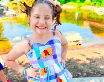 May include: A young girl wearing a blue and purple plaid dress with Winnie the Pooh characters printed on it. She has two Winnie the Pooh hair clips in her hair. She is smiling and looking at the camera.