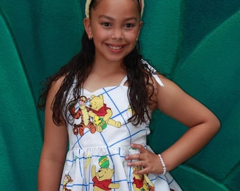 May include: A young girl wearing a white and blue dress with a Winnie the Pooh and Tigger print. She is wearing a yellow headband with bear ears and a blue denim skirt. The girl is standing in front of a green background.