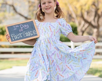 May include: A young girl smiles, holding a chalkboard that reads "1st Day of First Grade." She wears a light blue dress patterned with colorful pencils. The dress has short, puffy sleeves and a tiered skirt. The background is a blurred outdoor scene.