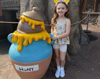 May include: A young girl stands next to a large, cartoonish honey pot. The pot is blue with a brown base and a brown lid, with yellow honey dripping down the sides. The pot has the word "HUNY" on it. The girl is wearing a Winnie the Pooh themed outfit and mouse ears.
