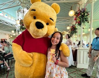 May include: A large, plush Winnie the Pooh character costume with yellow fur and a red shirt, standing next to a smiling child who is wearing a floral print top and holding a small Winnie the Pooh plush toy in a bright indoor setting.
