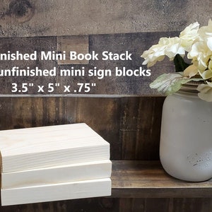May include: Three unfinished wooden blocks stacked on top of each other. The blocks are 3.5 inches by 5 inches by 0.75 inches. The blocks are on a wooden shelf with a white vase of white flowers on the right side of the shelf. The text "Unfinished Mini Book Stack" is above the blocks. The text "3 pack unfinished mini sign blocks" is below the blocks. The text "3.5" x 5" x .75" is below the text "3 pack unfinished mini sign blocks".