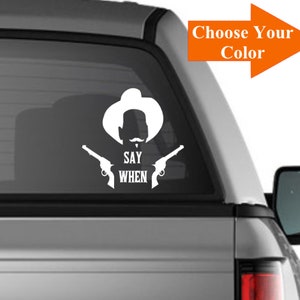May include: White vinyl decal of a cowboy with two pistols and the text "Say When". The decal is on the back window of a silver pickup truck.