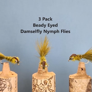 May include: Three Beady Eyed Damselfly Nymph Flies, displayed on corks against a blue background. The flies are yellow and brown, with detailed features. Text on the image reads "3 Pack Beady Eyed Damselfly Nymph Flies."