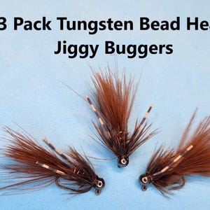 May include: Three brown and black Jiggy Bugger fishing flies with tungsten bead heads. Each fly has a dark hook and a tuft of brown fibers. The flies are arranged on a light blue background. The text "3 Pack Tungsten Bead Head Jiggy Buggers" is at the top.