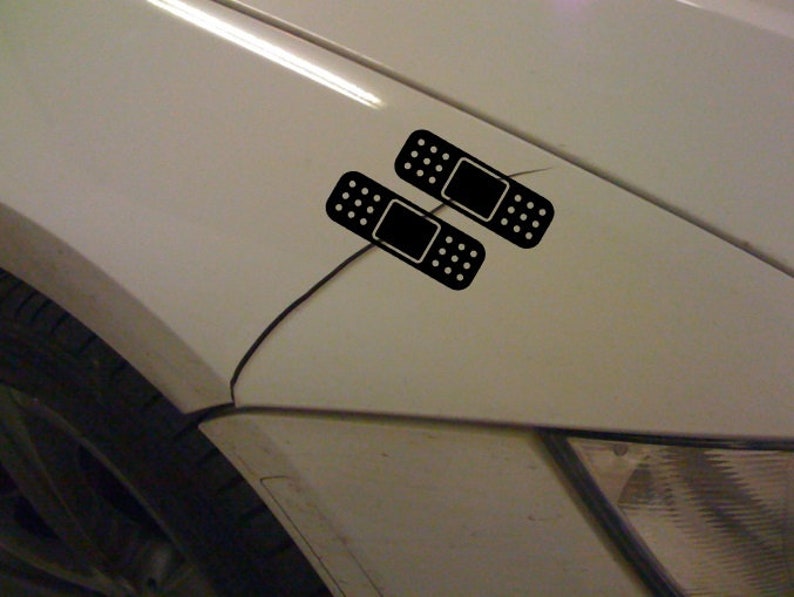 Puede incluir: Dos pegatinas de vendaje adhesivo negro en un guardabarros de coche blanco.