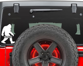 May include: White vinyl decal of a Bigfoot walking with the word "Believe" underneath. The decal is on the back window of a red Jeep.