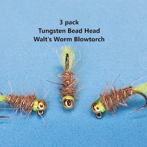 May include: Three fishing flies with gold tungsten bead heads, brown bodies, and chartreuse tails. The flies are arranged on a blue background. Text reads "3 pack Tungsten Bead Head Walt's Worm Blowtorch."