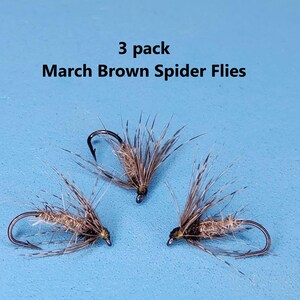 May include: Three March Brown Spider Flies on a blue background. Each fly has a dark hook, a brown body, and feathery wings. The text "3 pack March Brown Spider Flies" is above the flies.