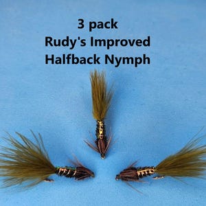 May include: Three Rudy's Improved Halfback Nymph fishing flies. Each fly has a dark body with gold accents and a tuft of olive-green fibers. The flies are arranged on a blue background, with the text "3 pack" and the fly name above them.