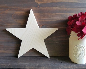 May include: A plain, unfinished wooden star with five points. The star is on a brown wooden background. A white painted mason jar with a floral design is to the right of the star. The jar is filled with red flowers.