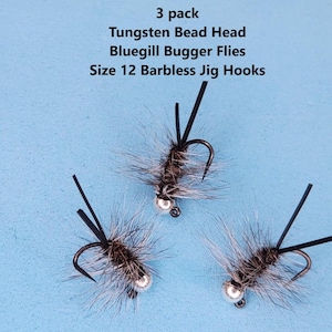 May include: Three Bluegill Bugger Flies with tungsten bead heads and size 12 barbless jig hooks. The flies have a silver bead head, black hook, and gray and brown body. The text in the image says "3 pack".