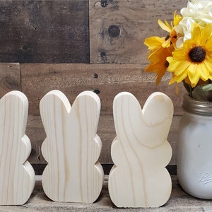 May include: Three unfinished wooden bunny shapes, standing in a row on a woodgrain background. A white mason jar with yellow and white flowers is to the right of the bunnies.