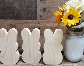 May include: Three unfinished wooden bunny shapes, standing in a row on a woodgrain background. A white mason jar with yellow and white flowers is to the right of the bunnies.