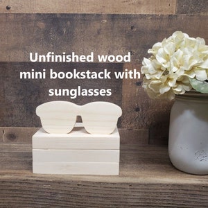 May include: Unfinished wood mini bookstack with sunglasses. The image shows a stack of three rectangular wooden blocks with a wooden sunglasses cutout on top. The text "Unfinished wood mini bookstack with sunglasses" is displayed in white.