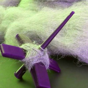 Peut inclure: Une roue &agrave; filer violette avec une broche et une pelote de fil gris clair. La roue &agrave; filer est sur une surface verte.