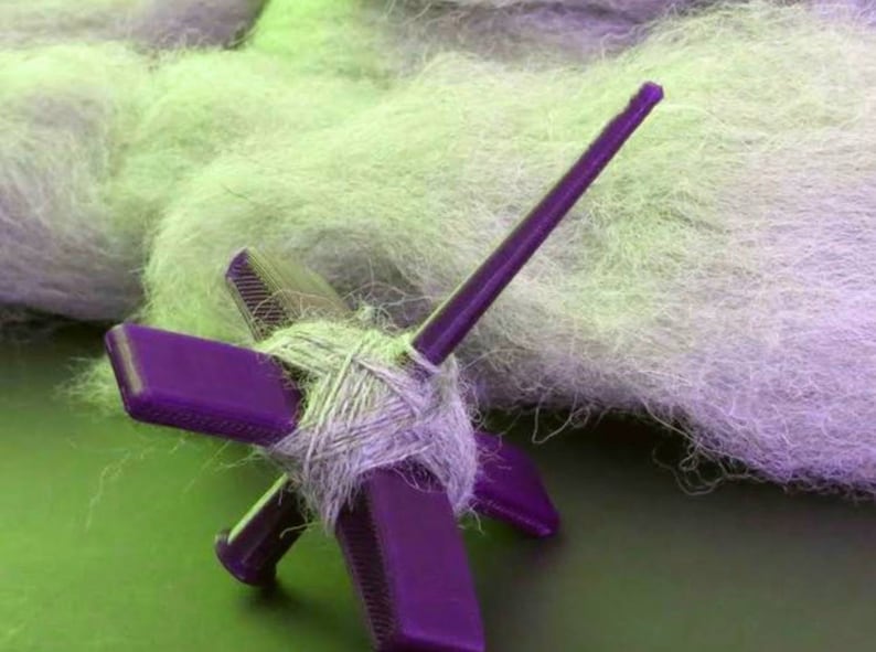 Peut inclure: Une roue &agrave; filer violette avec une broche et une pelote de fil gris clair. La roue &agrave; filer est sur une surface verte.