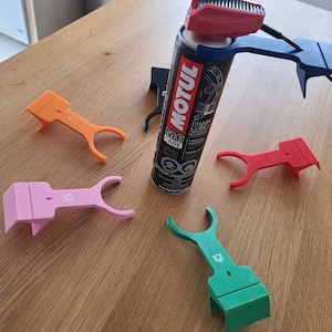 Pu&ograve; includere: Cinque porta-bottiglie spray in plastica stampati in 3D, in arancione, rosa, rosso, blu e verde. I porta-bottiglie sono progettati per mantenere una bottiglia spray in posizione verticale. Una bottiglia spray di lubrificante per catena Motul MC Care &egrave; al centro dell'immagine.