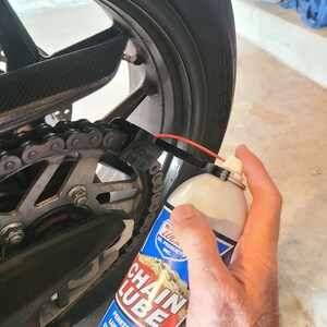 Pu&ograve; includere: Una bomboletta di lubrificante per catena rossa e bianca con le parole "Chain Lube" in grandi caratteri grassetto. La bomboletta viene spruzzata su una catena di moto.