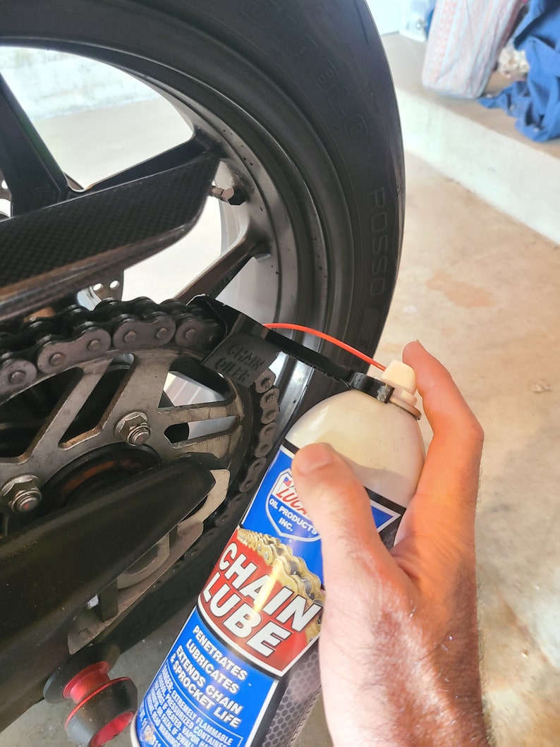 Pu&ograve; includere: Una bomboletta di lubrificante per catena rossa e bianca con le parole "Chain Lube" in grandi caratteri grassetto. La bomboletta viene spruzzata su una catena di moto.