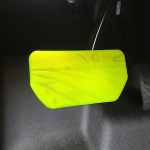 May include: A bright neon yellow car brake pedal cover featuring a textured surface with subtle geometric lines and a circular indentation. The rectangular pedal cover is constructed with a smooth finish and is mounted within a dark vehicle interior.