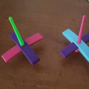 Peut inclure: Deux toupies en bois color&eacute;es. Une toupie a un sch&eacute;ma de couleurs violet, rose et vert. L'autre toupie a un sch&eacute;ma de couleurs violet, bleu et rose.