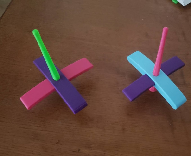 Peut inclure: Deux toupies en bois color&eacute;es. Une toupie a un sch&eacute;ma de couleurs violet, rose et vert. L'autre toupie a un sch&eacute;ma de couleurs violet, bleu et rose.