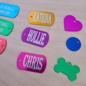 May include: Assortment of colorful metal dog tags with engraved names, including "Katrina", "Hollie", "Chris", and "Get My Ballz Since 2021".  Also included are a green bone-shaped tag, a pink heart-shaped tag, and a blue circle tag.