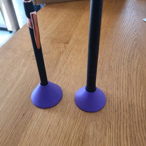 May include: Two black digital styluses standing upright in purple cone-shaped holders. One stylus features a rose gold clip and accents, while the other is solid black with a textured grip area. Both pens are positioned vertically on their bases.