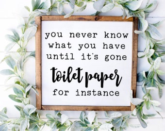 May include: A framed wooden sign with the text "you never know what you have until it's gone toilet paper for instance." The sign is surrounded by a garland of green and white leaves.