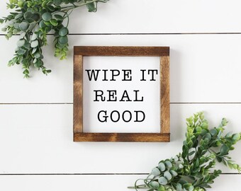 May include: A square wooden sign with the phrase "WIPE IT REAL GOOD" in black letters. The sign has a dark brown frame and is set against a white background. Greenery is placed around the sign.