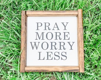 May include: A square wooden sign with the words "PRAY MORE WORRY LESS" in gray lettering. The sign has a white background and a dark brown wooden frame. The sign is set on a bed of green grass.