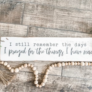 May include: A white wooden sign with the quote "I still remember the days I prayed for the things I have now" in black lettering. The sign is decorated with greenery and a wooden bead garland.