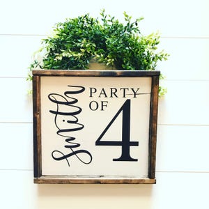 May include: A wooden framed sign with the text "Smith Party of 4" in black lettering. The sign has a white background and is decorated with a green wreath on top.
