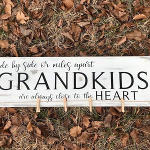 May include: A rectangular, distressed white wooden sign with the words "Side by Side or miles apart GRANDKIDS are always close to the HEART" in black script. Small wooden clothespins are attached to a string across the bottom.